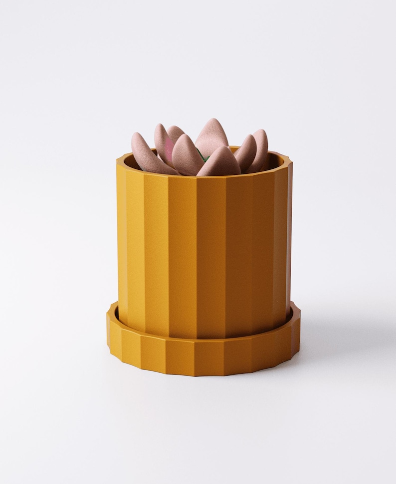 Puede incluir: Una maceta cil&iacute;ndrica de color amarillo mostaza con estr&iacute;as verticales y una base de plato circular a juego. Dentro del recipiente hay una peque&ntilde;a planta suculenta estilizada con hojas gruesas, redondeadas y de color rosa p&aacute;lido.