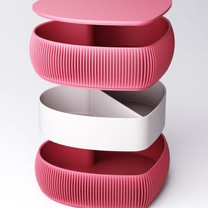May include: A pink heart-shaped storage container with a lid, a white interior compartment, and a ribbed exterior. The lid has a small circular opening. The container is designed to be stacked.