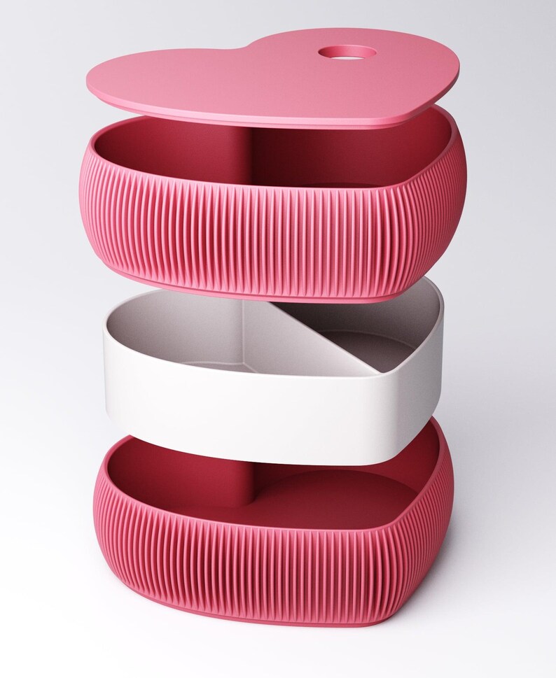 May include: A pink heart-shaped storage container with a lid, a white interior compartment, and a ribbed exterior. The lid has a small circular opening. The container is designed to be stacked.