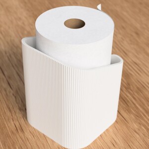 Op de afbeelding: Een witte toiletpapierrol in een bijpassende houder. De houder heeft een gebogen ontwerp met verticale lijnen. De achtergrond is een lichtbruin houten oppervlak. De toiletpapierrol is wit.