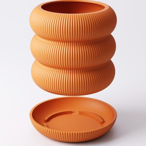 May include: A terracotta-coloured, ribbed ceramic plant pot with a matching saucer. The pot has a distinctive stacked design, with three rounded sections. The saucer is round with a raised edge and a simple design. The set is a warm, earthy tone.