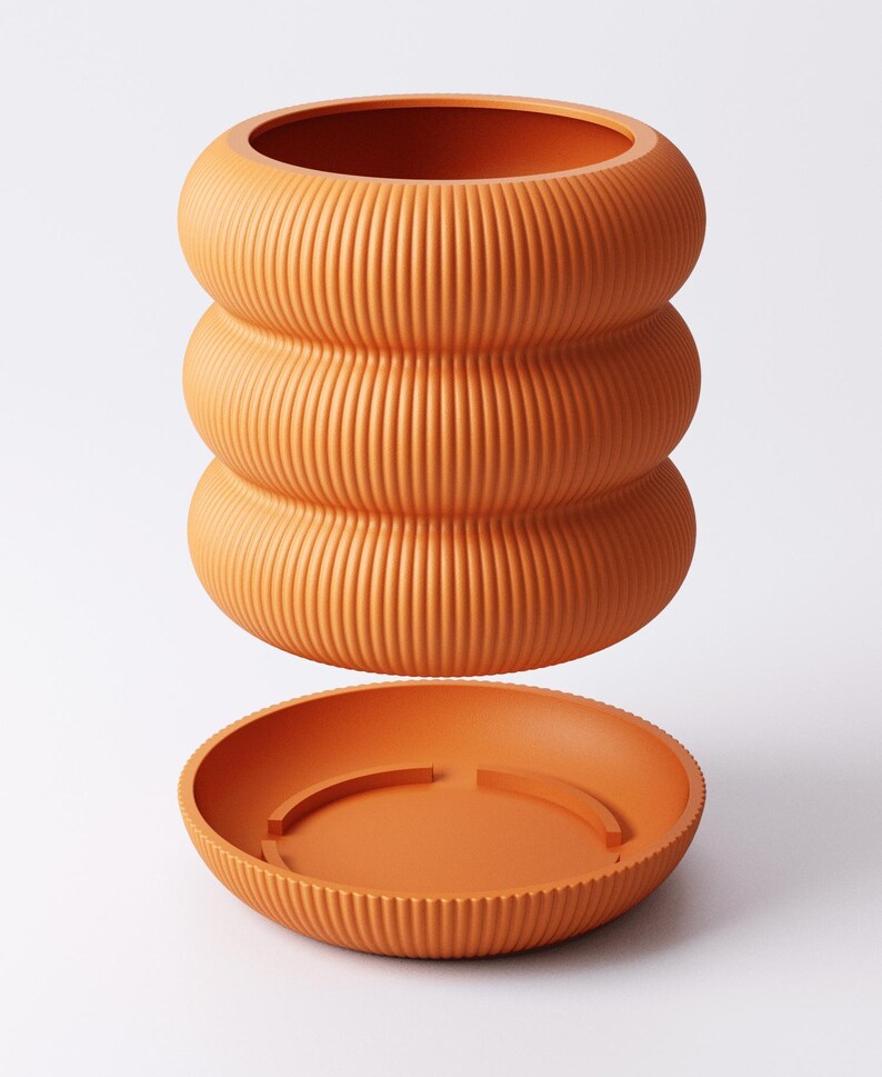 May include: A terracotta-coloured, ribbed ceramic plant pot with a matching saucer. The pot has a distinctive stacked design, with three rounded sections. The saucer is round with a raised edge and a simple design. The set is a warm, earthy tone.