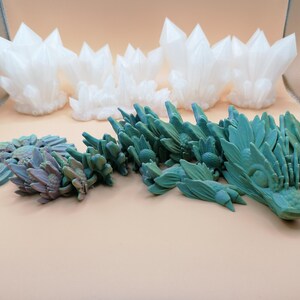 May include: An assortment of 3D-printed fantasy sculptures. The collection includes white crystal formations and a teal-colored dragon-like creature. A smaller, flower-like sculpture is also present, with a mix of teal and purple hues.