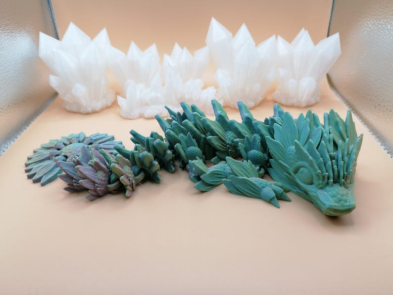 May include: An assortment of 3D-printed fantasy sculptures. The collection includes white crystal formations and a teal-colored dragon-like creature. A smaller, flower-like sculpture is also present, with a mix of teal and purple hues.