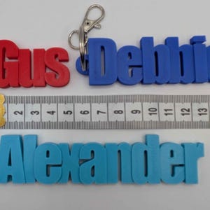 May include: Three personalized keychains in red, blue, and light blue, spelling out "Gus", "Debbie", and "Alexander". A ruler shows measurements in inches. Each keychain has a metal clasp.