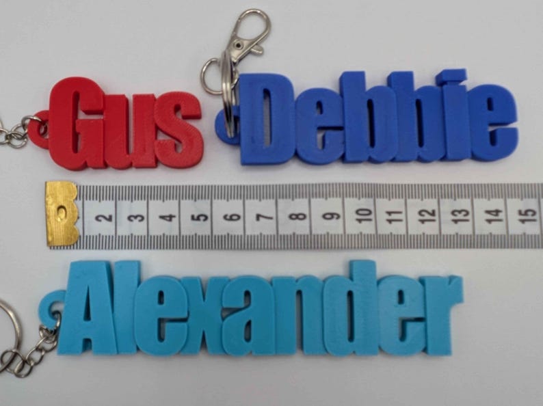 May include: Three personalized keychains in red, blue, and light blue, spelling out "Gus", "Debbie", and "Alexander". A ruler shows measurements in inches. Each keychain has a metal clasp.