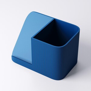 May include: A two-toned blue desk organizer with a rectangular compartment and a slanted section. The rectangular section has vertical ridged detailing. The organizer is made of a solid material and sits on a white surface.