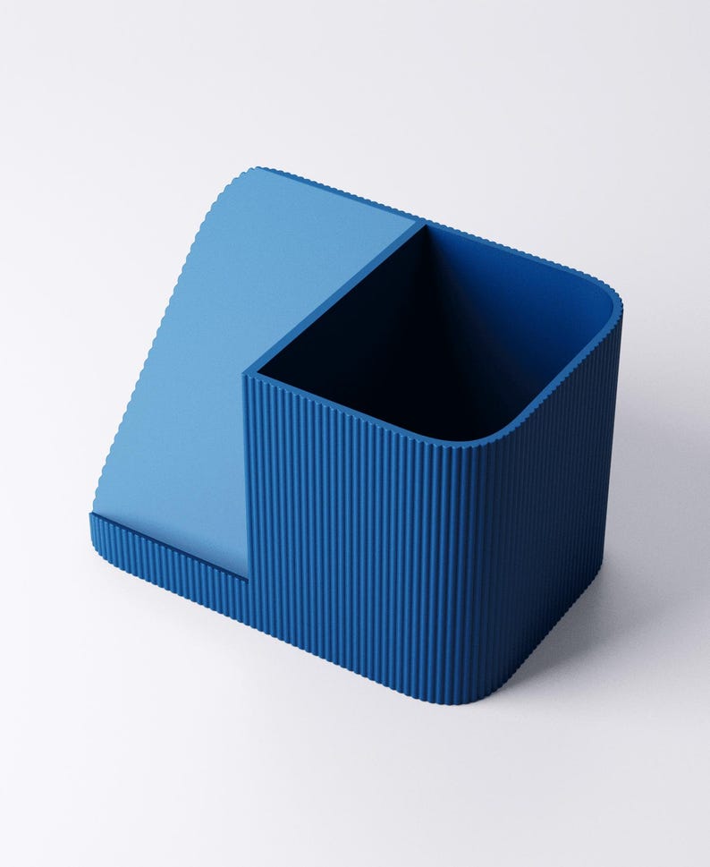 May include: A two-toned blue desk organizer with a rectangular compartment and a slanted section. The rectangular section has vertical ridged detailing. The organizer is made of a solid material and sits on a white surface.