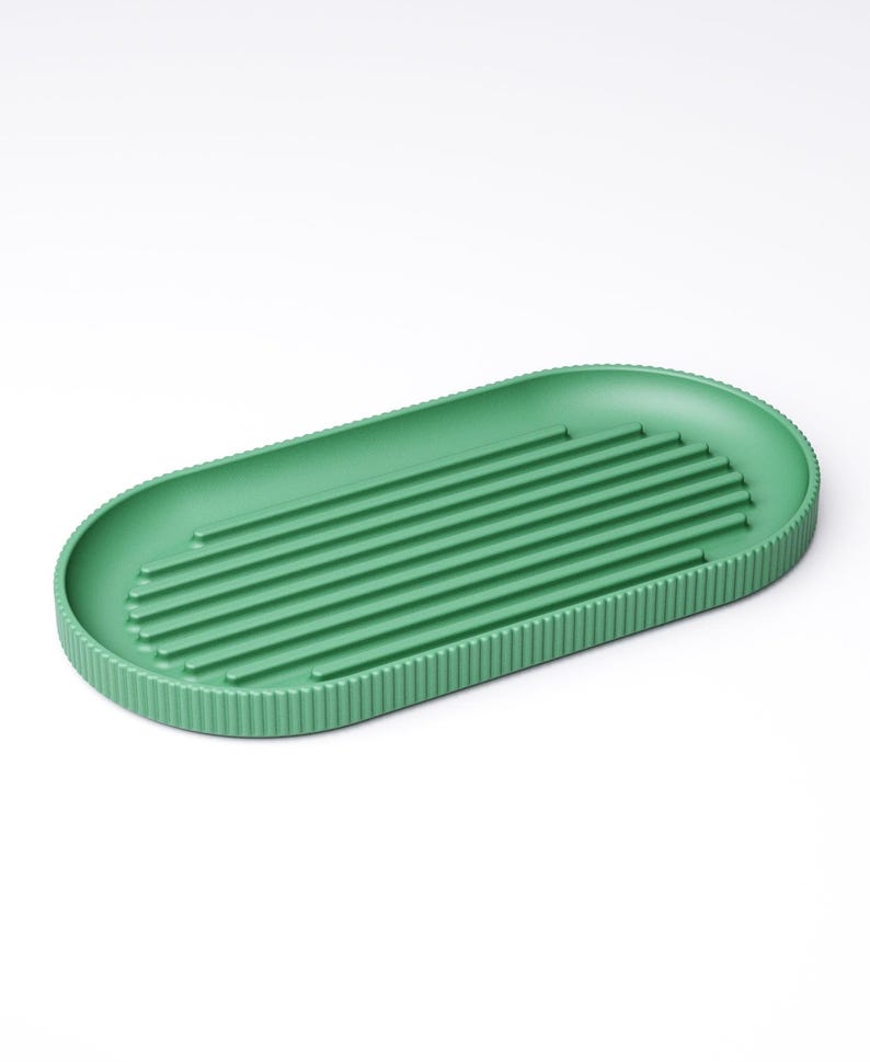 May include: A green, oval-shaped soap dish with a ribbed design. The dish has a series of parallel ridges across the center for drainage and a textured border. The soap dish is made of a solid, matte material.