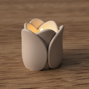 May include: A beige tulip-shaped candle holder with a lit candle inside. The holder has a textured surface and is sitting on a wooden surface. The candle holder is designed to resemble a tulip flower.