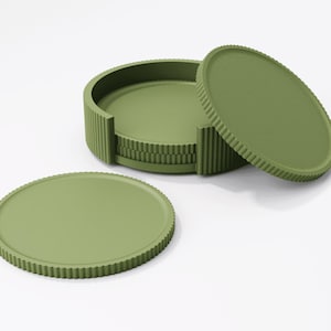 May include: A set of olive green coasters with a ridged edge design. The set includes a holder and several individual coasters, all in a matching muted green color. The coasters are round and appear to be made of a solid material.