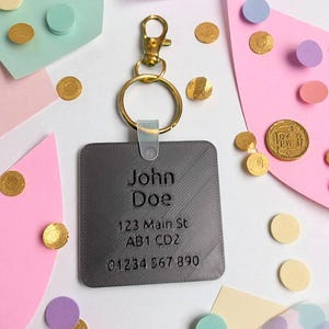 May include: A gray rectangular keychain tag with the engraved name "John Doe", address, and phone number. The tag is attached to a gold-colored clasp and ring. The background features colorful paper shapes and gold confetti.