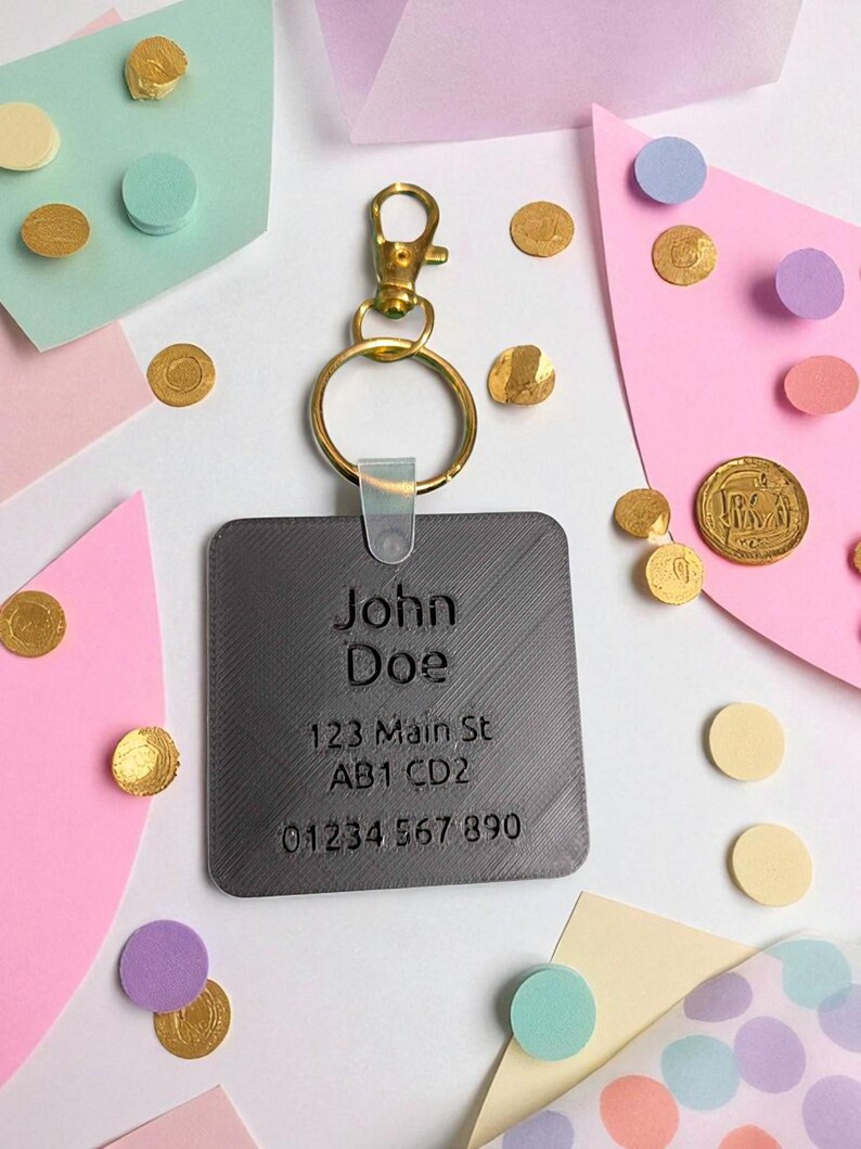 May include: A gray rectangular keychain tag with the engraved name "John Doe", address, and phone number. The tag is attached to a gold-colored clasp and ring. The background features colorful paper shapes and gold confetti.