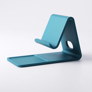 May include: A teal-colored phone stand with a minimalist design. The stand features a rectangular base, a curved support, and a small circular hole. The stand is designed to hold a phone or tablet.