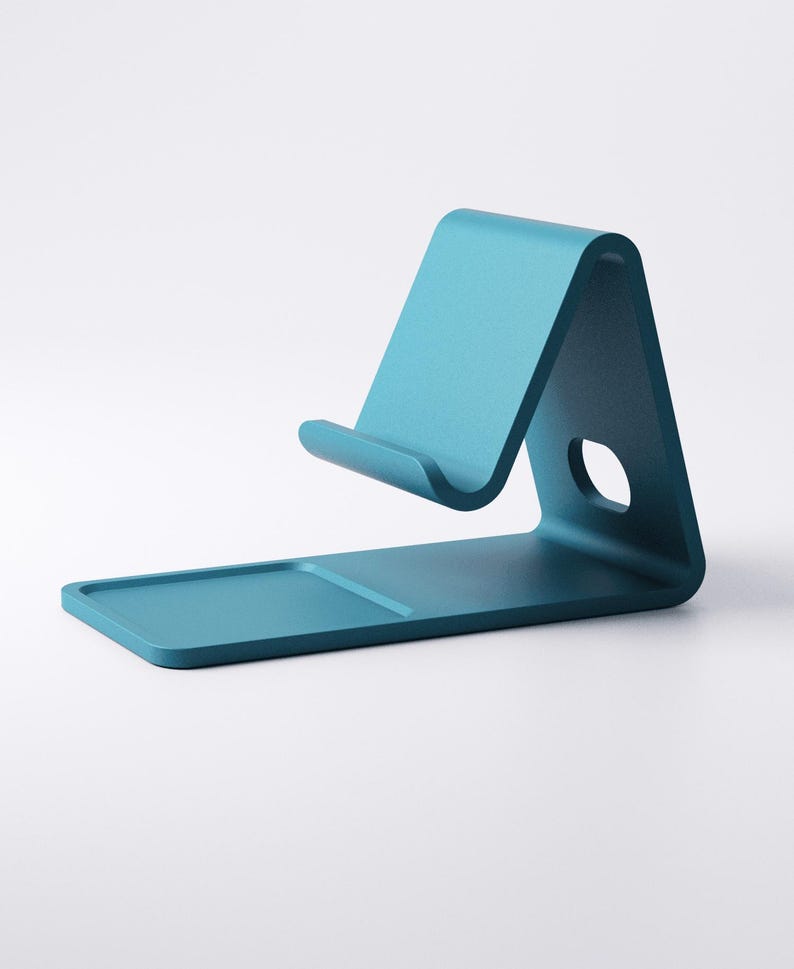 May include: A teal-colored phone stand with a minimalist design. The stand features a rectangular base, a curved support, and a small circular hole. The stand is designed to hold a phone or tablet.