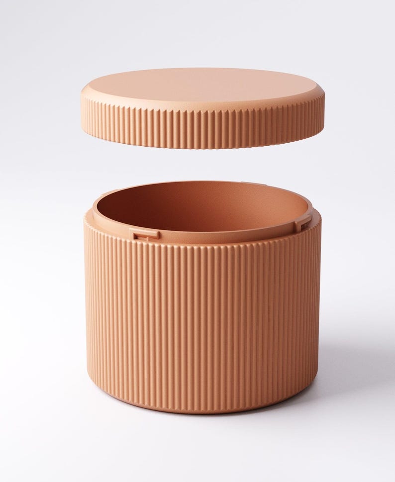 May include: A peach-colored, cylindrical container with a matching lid. The lid is slightly elevated, revealing the interior of the container. Both the lid and the container have vertical, ridged detailing, creating a textured appearance. The container is designed to store items.