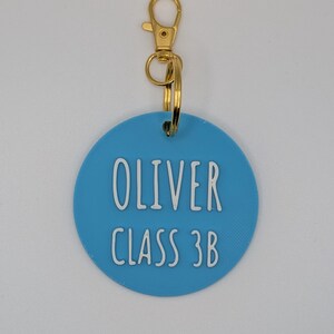 May include: A light blue, circular keychain tag with the name "OLIVER" and "CLASS 3B" in white lettering. The tag is attached to a gold-colored clasp and ring.