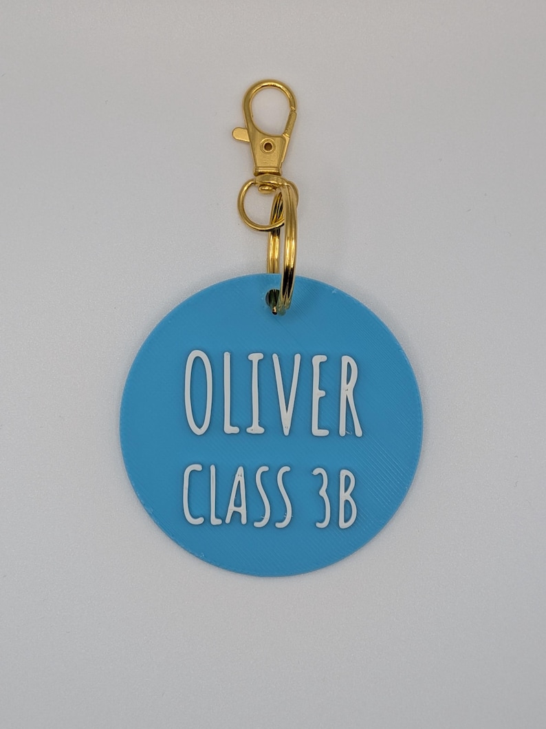 May include: A light blue, circular keychain tag with the name "OLIVER" and "CLASS 3B" in white lettering. The tag is attached to a gold-colored clasp and ring.