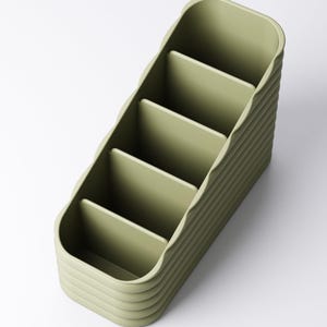 May include: A sage green desk organizer with five compartments. The organizer has a ribbed exterior design and a curved shape. The compartments are open and ready to hold pens, pencils, or other office supplies. The organizer is made of a solid material.