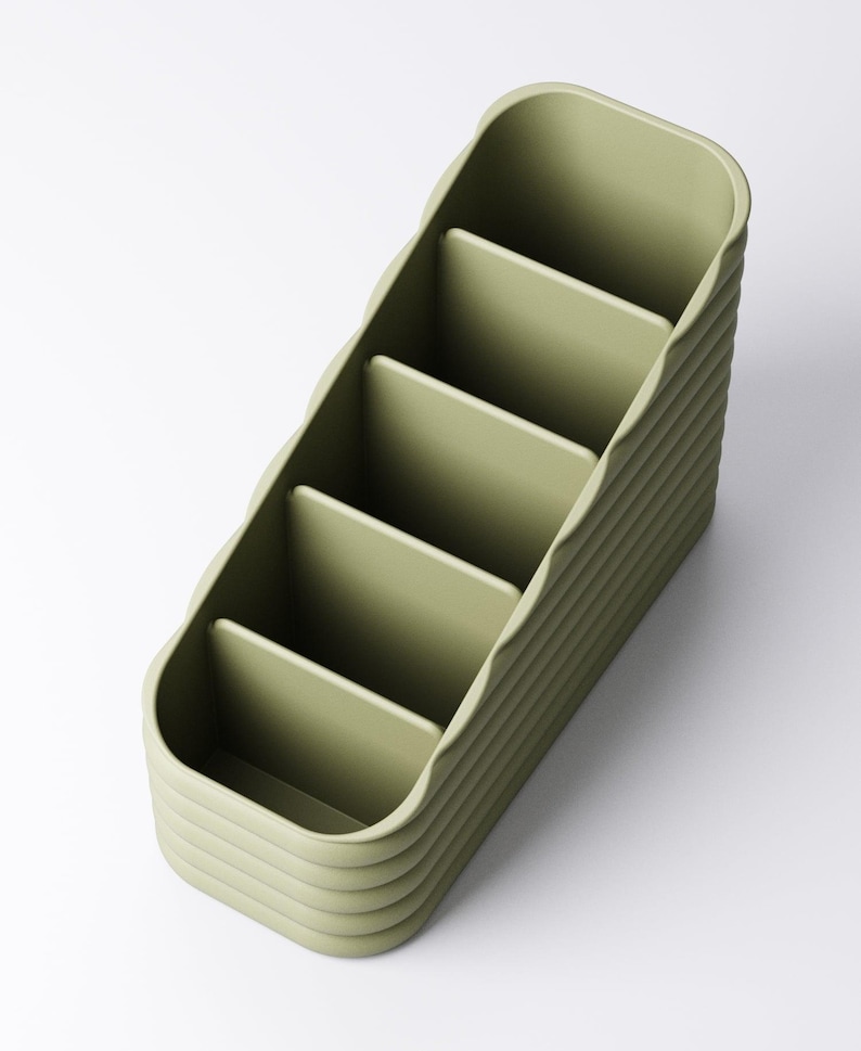 May include: A sage green desk organizer with five compartments. The organizer has a ribbed exterior design and a curved shape. The compartments are open and ready to hold pens, pencils, or other office supplies. The organizer is made of a solid material.