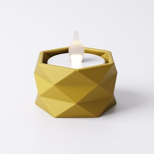 May include: A geometric, mustard-yellow candle holder with a white, glowing LED flame. The holder has a faceted design and a hexagonal shape. The flame is shaped like a teardrop and emits a soft, warm light, creating a modern, decorative accent.