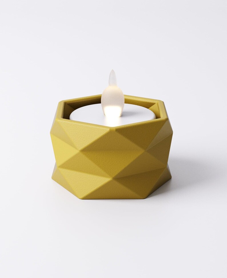 May include: A geometric, mustard-yellow candle holder with a white, glowing LED flame. The holder has a faceted design and a hexagonal shape. The flame is shaped like a teardrop and emits a soft, warm light, creating a modern, decorative accent.
