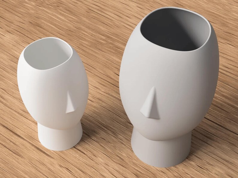 May include: Two white ceramic head-shaped vases. The vases have a smooth, matte finish and a simple, minimalist design. The larger vase is approximately 10 inches tall, while the smaller one is about 6 inches tall. The vases are placed on a wooden surface.