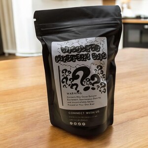Op de afbeelding: Een zwarte zak met ritssluiting, met het label "Wiggles Mystery Bag". Het witte label heeft een groot vraagteken en kleinere vraagtekens. De tekst luidt "Waarschuwing: Inhoud kan extreme opwinding veroorzaken..."