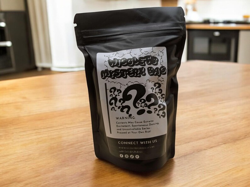 Op de afbeelding: Een zwarte zak met ritssluiting, met het label "Wiggles Mystery Bag". Het witte label heeft een groot vraagteken en kleinere vraagtekens. De tekst luidt "Waarschuwing: Inhoud kan extreme opwinding veroorzaken..."
