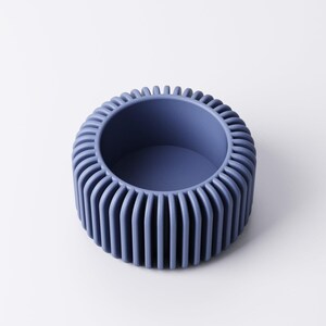 May include: A round, blue decorative bowl with a ribbed exterior. The bowl has a smooth, matte finish and a circular opening. The ribbed design creates a textured surface. The bowl is a solid color and sits on a white surface.