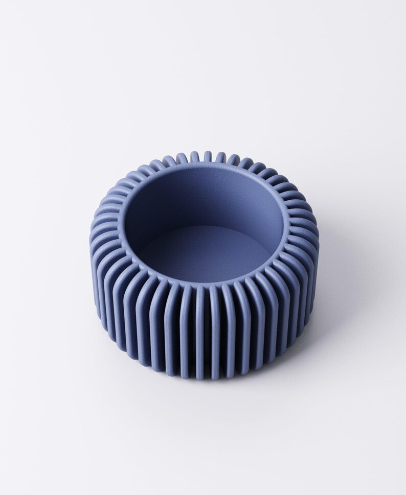 May include: A round, blue decorative bowl with a ribbed exterior. The bowl has a smooth, matte finish and a circular opening. The ribbed design creates a textured surface. The bowl is a solid color and sits on a white surface.