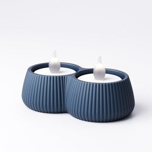 May include: Two blue candle holders with a ribbed design, each holding a white LED candle with a flickering flame. The holders are connected, creating a double candle display. The background is plain white.