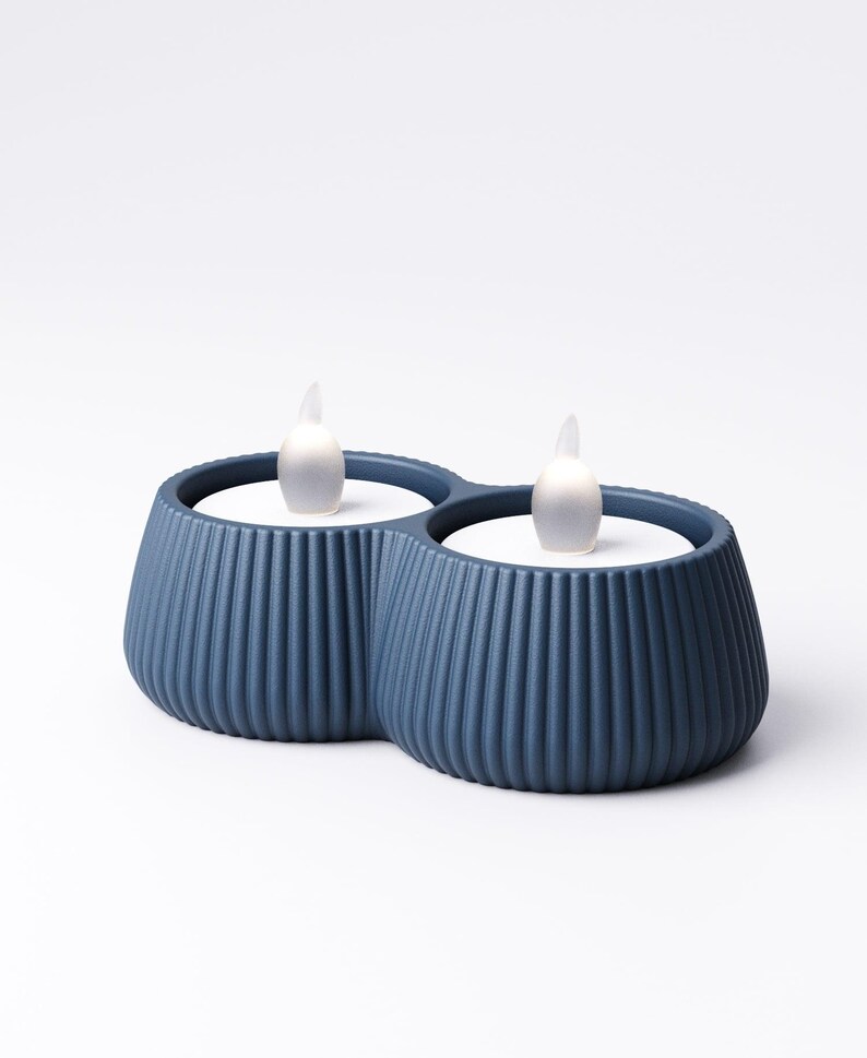 May include: Two blue candle holders with a ribbed design, each holding a white LED candle with a flickering flame. The holders are connected, creating a double candle display. The background is plain white.
