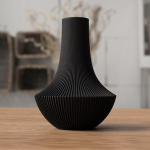 May include: A black decorative vase with a unique, ribbed design. The vase has a wide, rounded base that tapers upwards to a narrower neck. The vase is made of a solid, dark material and sits on a wooden surface. The background is blurred, suggesting a home decor setting.