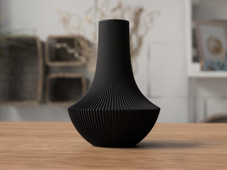 May include: A black decorative vase with a unique, ribbed design. The vase has a wide, rounded base that tapers upwards to a narrower neck. The vase is made of a solid, dark material and sits on a wooden surface. The background is blurred, suggesting a home decor setting.