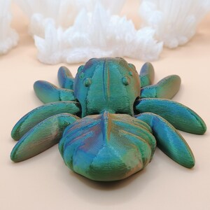 Op de afbeelding: Een 3D-geprinte iriserende spinnenfiguur in tinten teal, blauw en groen. De spin heeft acht poten en een getextureerd lichaam. Witte, kristallijne formaties staan op de achtergrond, tegen een lichtbeige oppervlak.