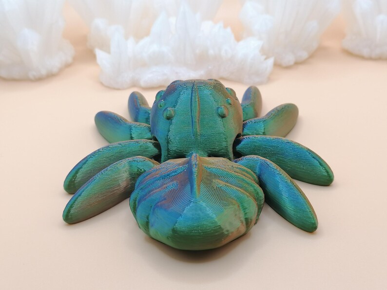 Op de afbeelding: Een 3D-geprinte iriserende spinnenfiguur in tinten teal, blauw en groen. De spin heeft acht poten en een getextureerd lichaam. Witte, kristallijne formaties staan op de achtergrond, tegen een lichtbeige oppervlak.