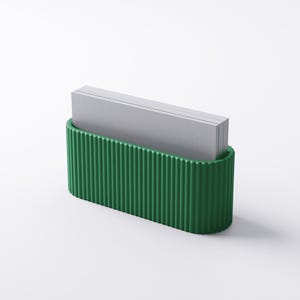 May include: A green, ribbed business card holder with a curved base. The holder contains a stack of gray business cards. The design is minimalist and modern, suitable for an office or desk setting.