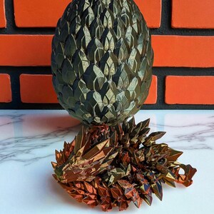 Op de afbeelding: Een bronzen draaksculptuur en een bijpassend ei. Het ei is donkergroen en heeft een schubbenpatroon. De draak ligt en heeft een gedetailleerd, geometrisch ontwerp. Bakstenen muur op de achtergrond.