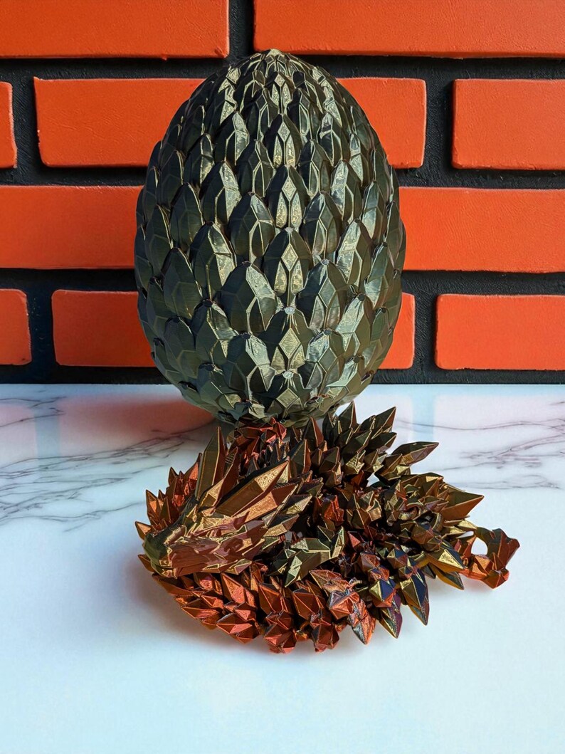 Op de afbeelding: Een bronzen draaksculptuur en een bijpassend ei. Het ei is donkergroen en heeft een schubbenpatroon. De draak ligt en heeft een gedetailleerd, geometrisch ontwerp. Bakstenen muur op de achtergrond.