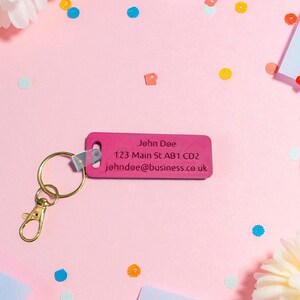 May include: A pink rectangular keychain with gold hardware and the text "John Doe" and contact information. The keychain is on a pink surface with confetti and white paper notes.