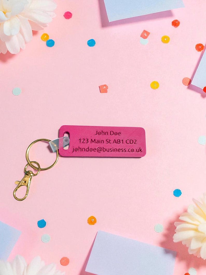 May include: A pink rectangular keychain with gold hardware and the text "John Doe" and contact information. The keychain is on a pink surface with confetti and white paper notes.