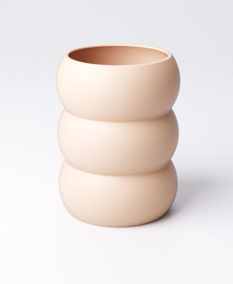May include: A peach-colored ceramic vase with a unique stacked design. The vase is composed of three rounded sections, creating a bulbous shape. The top section has a wide opening. The smooth, matte finish gives it a modern aesthetic. Ideal for home decor.