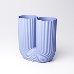 K&ouml;nnte beinhalten: Eine hell-lavendelblaue U-f&ouml;rmige Vase mit einer durchgehenden vertikalen Rippenstruktur auf der gesamten Au&szlig;enfl&auml;che. Das Objekt verf&uuml;gt &uuml;ber zwei offene zylindrische &Ouml;ffnungen an der Oberseite und einen abgerundeten Boden, was eine symmetrische Doppelform ergibt.