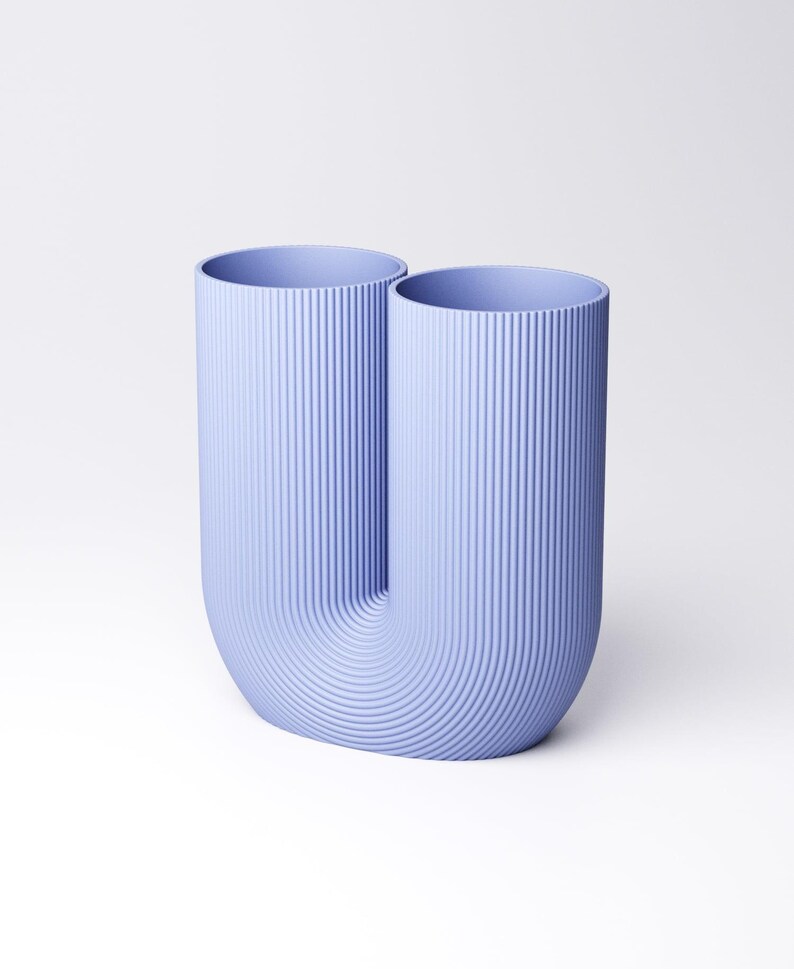 K&ouml;nnte beinhalten: Eine hell-lavendelblaue U-f&ouml;rmige Vase mit einer durchgehenden vertikalen Rippenstruktur auf der gesamten Au&szlig;enfl&auml;che. Das Objekt verf&uuml;gt &uuml;ber zwei offene zylindrische &Ouml;ffnungen an der Oberseite und einen abgerundeten Boden, was eine symmetrische Doppelform ergibt.