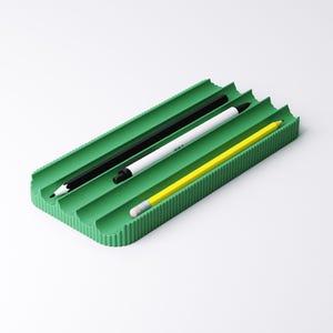 May include: A green rectangular desk organizer with three slots, holding a black pencil, a white pen, and a yellow pencil. The organizer has a ribbed texture and is set against a white background.