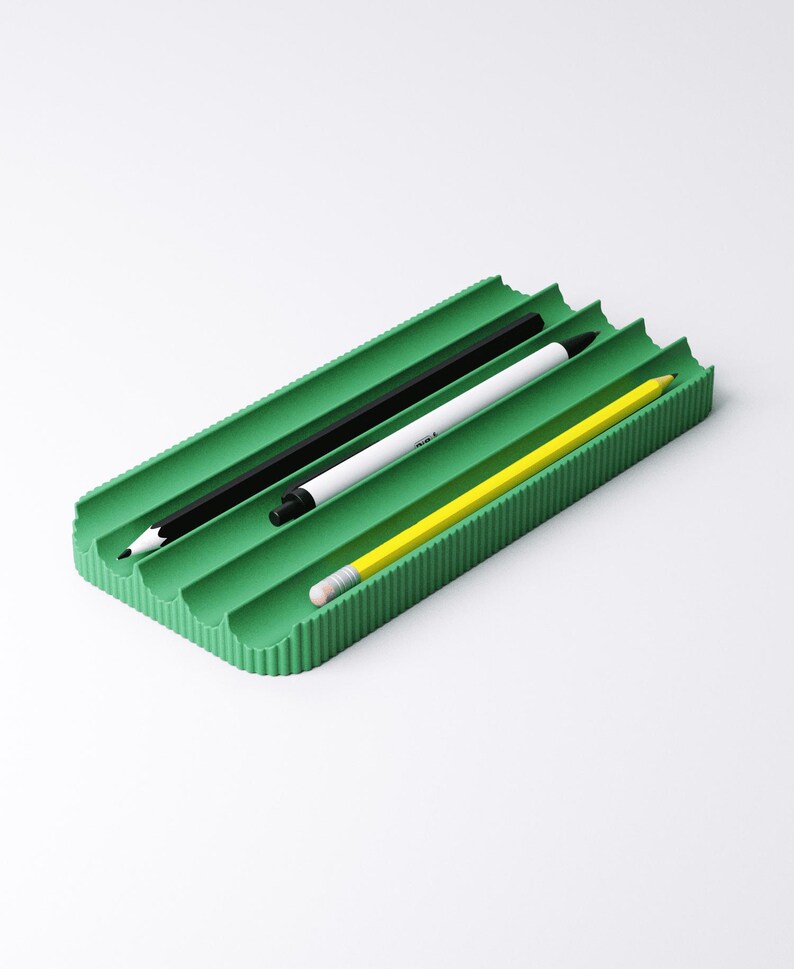 May include: A green rectangular desk organizer with three slots, holding a black pencil, a white pen, and a yellow pencil. The organizer has a ribbed texture and is set against a white background.