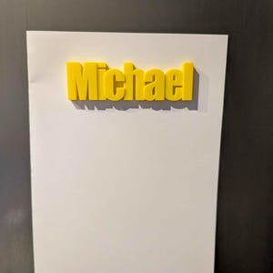 May include: A yellow 3D-printed name, "Michael", is affixed to a white sheet of paper. The paper is attached to a dark surface, likely a refrigerator or metal board. The name is in a bold, sans-serif font.