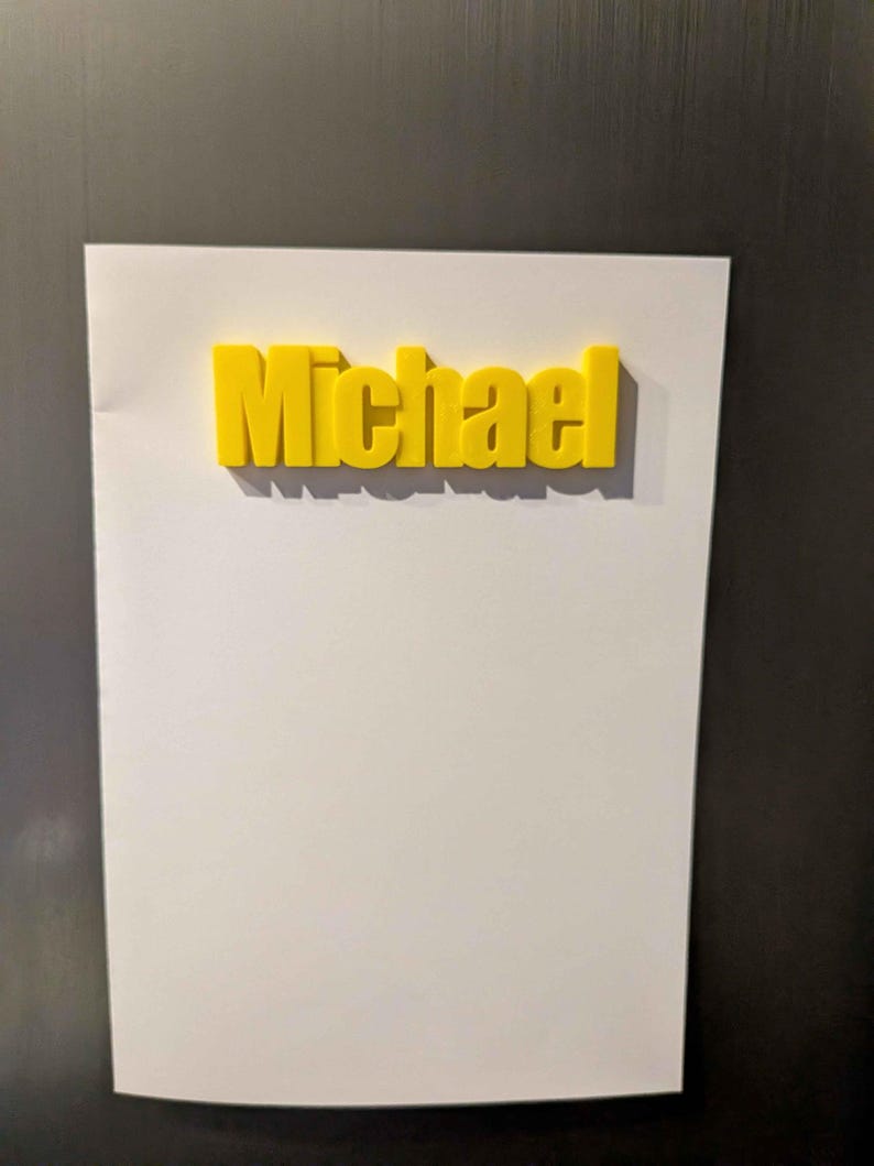 May include: A yellow 3D-printed name, "Michael", is affixed to a white sheet of paper. The paper is attached to a dark surface, likely a refrigerator or metal board. The name is in a bold, sans-serif font.