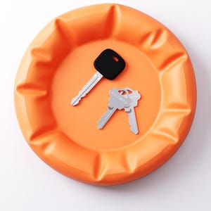 May include: An orange, round catch-all tray with a ridged edge. Two keys rest inside the tray: one with a black head and the other two with silver heads. The keys are likely for a house or car.